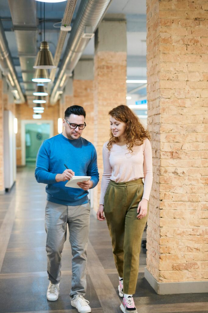 Two business colleagues walking in an office hallway discussing ideas with a notepad.